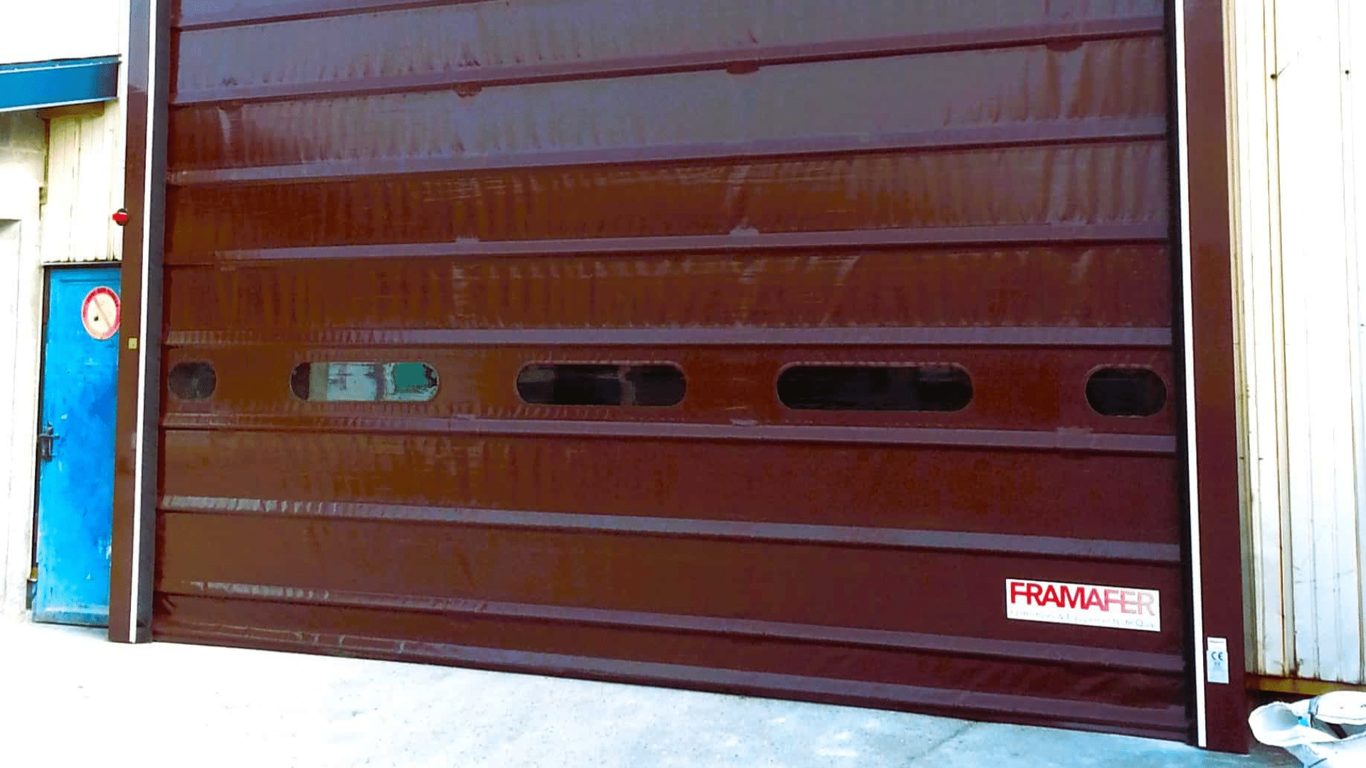 Porte industrielle en PVC de couleur marron, équipée de fenêtres rectangulaires, installée dans un bâtiment avec un logo 'FRAMAFER' visible en bas à droite